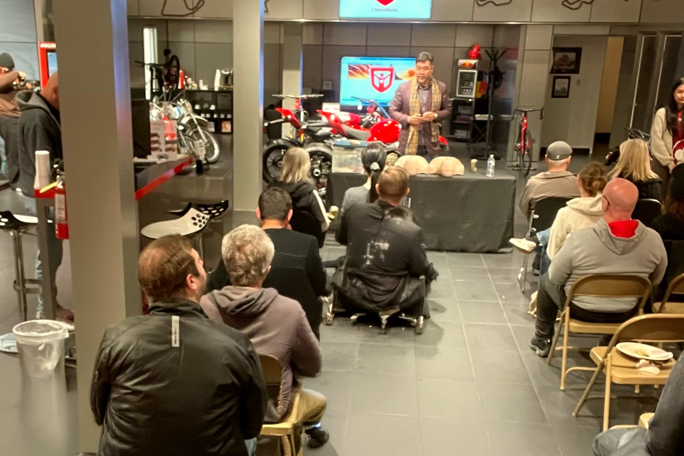 Kai Chang teaching a Citizen Medic workshop to a seated group of motorcyclists, CPR mannequins on the table in front of him